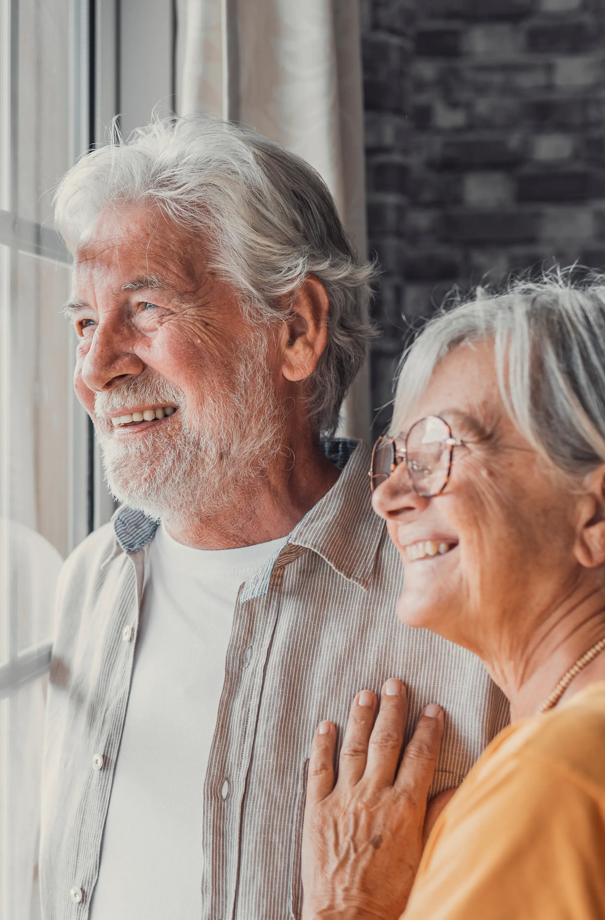 Happy bonding loving middle aged senior retired couple standing near window, looking in distance, recollecting good memories or planning common future, enjoying peaceful moment together at home